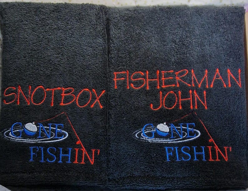 May include: Two black rectangular towels featuring embroidered text and graphics. The left towel reads SNOTBOX and the right reads FISHERMAN JOHN in red. Both feature a blue and white fishing bobber graphic with the text GONE FISHIN' in blue.