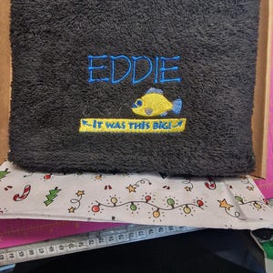 May include: A dark gray rectangular towel featuring embroidered text that reads EDDIE in blue and IT WAS THIS BIG! in yellow, accompanied by a yellow fish graphic. A white measuring tape is visible below, showing measurements in centimeters.