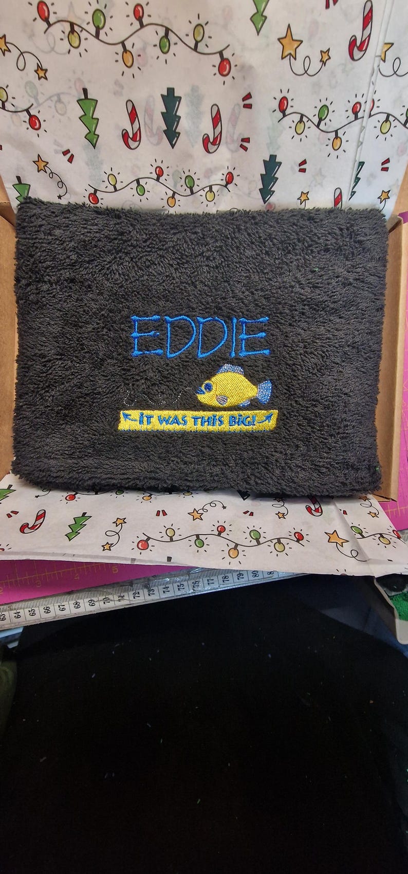 May include: A dark gray rectangular towel featuring embroidered text that reads EDDIE in blue and IT WAS THIS BIG! in yellow, accompanied by a yellow fish graphic. A white measuring tape is visible below, showing measurements in centimeters.