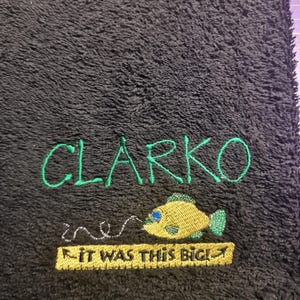 May include: A black textured towel featuring green embroidered text that reads CLARKO. Below the name is a yellow fish with blue accents and a silver fishing line, followed by a yellow rectangular patch with the text IT WAS THIS BIG!