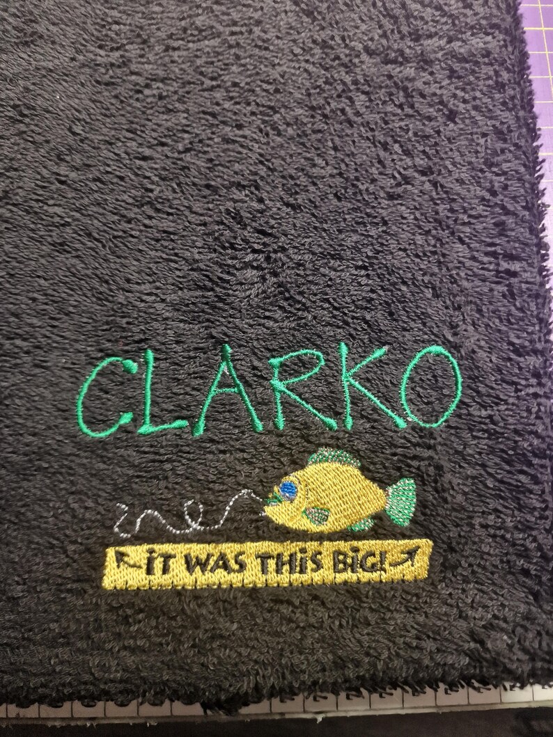 May include: A black textured towel featuring green embroidered text that reads CLARKO. Below the name is a yellow fish with blue accents and a silver fishing line, followed by a yellow rectangular patch with the text IT WAS THIS BIG!