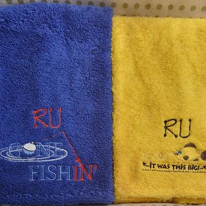 May include: A blue towel with embroidered text RU GONE FISHIN' and a fishing bobber graphic, alongside a yellow towel with embroidered text RU IT WAS THIS BIG! and a fish graphic. Both towels feature a soft, textured terry cloth fabric.