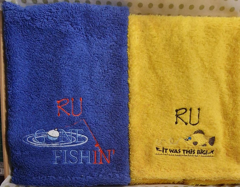 May include: A blue towel with embroidered text RU GONE FISHIN' and a fishing bobber graphic, alongside a yellow towel with embroidered text RU IT WAS THIS BIG! and a fish graphic. Both towels feature a soft, textured terry cloth fabric.