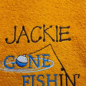 May include: A bright orange towel featuring black and blue embroidery. The text reads JACKIE GONE FISHIN' in a mix of black and blue lettering, accompanied by a stitched graphic of a fishing rod and a blue and white bobber.