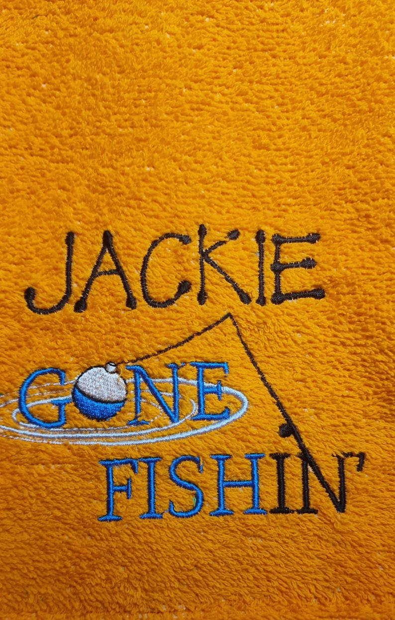 May include: A bright orange towel featuring black and blue embroidery. The text reads JACKIE GONE FISHIN' in a mix of black and blue lettering, accompanied by a stitched graphic of a fishing rod and a blue and white bobber.