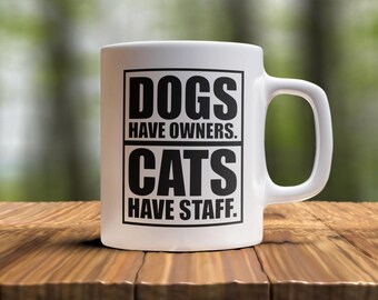 May include: White ceramic coffee mug with black text that reads "Dogs have owners. Cats have staff."