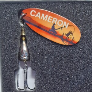 May include: A fishing lure featuring an oval orange blade with the name CAMERON printed in white, alongside a graphic of a person fishing and a fish. The lure includes a metallic silver body and a triple hook attached to the bottom.
