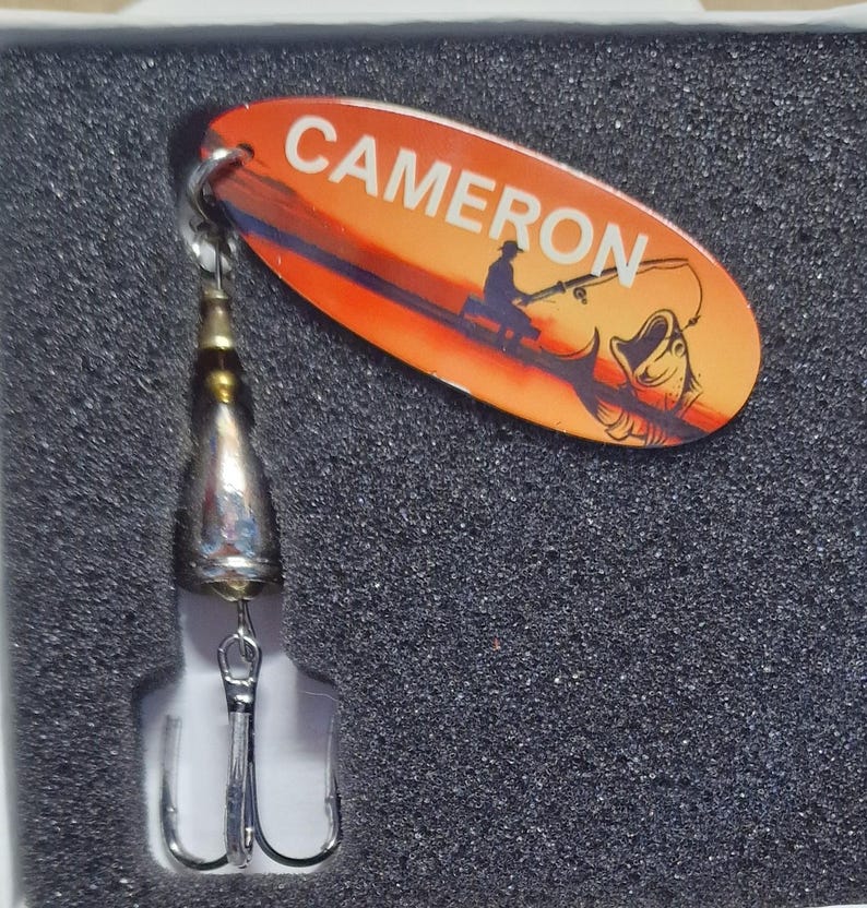 May include: A fishing lure featuring an oval orange blade with the name CAMERON printed in white, alongside a graphic of a person fishing and a fish. The lure includes a metallic silver body and a triple hook attached to the bottom.