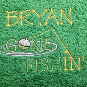 May include: A bright green towel featuring embroidered text that reads BRYAN GONE FISHIN'. The design includes a yellow fishing rod, a green and white bobber floating in water ripples, and yellow lettering for the name and the word FISHIN'.