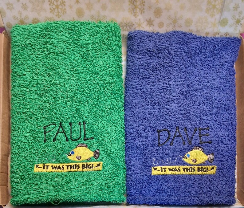 May include: Two rectangular terry cloth towels, one green and one blue. The green towel is embroidered with the name Paul, a yellow fish, and the text IT WAS THIS BIG! on a yellow ruler. The blue towel features the name Dave, a yellow fish, and the same text.