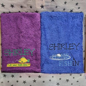 May include: Two rectangular terry cloth towels, one purple and one blue. The purple towel features green embroidery reading SHIRLEY and a yellow fish with the text IT WAS THIS BIG. The blue towel has black embroidery reading SHIRLEY GONE FISHIN.