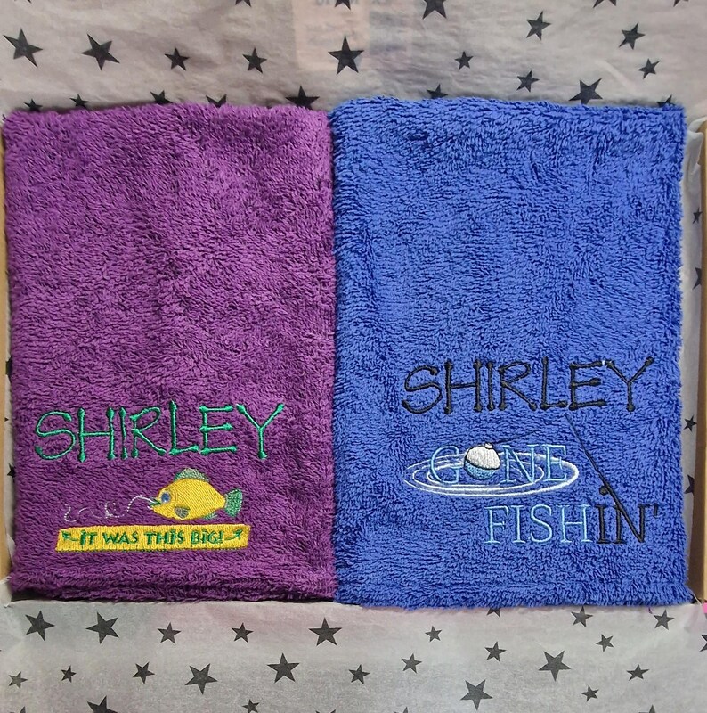 May include: Two rectangular terry cloth towels, one purple and one blue. The purple towel features green embroidery reading SHIRLEY and a yellow fish with the text IT WAS THIS BIG. The blue towel has black embroidery reading SHIRLEY GONE FISHIN.