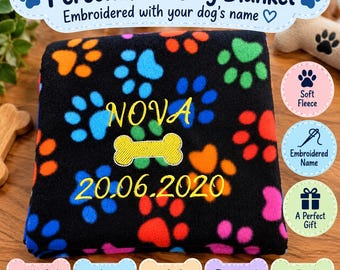May include: A black fleece blanket featuring a colorful pattern of paw prints in orange, blue, green, red, and pink. The blanket is embroidered with the name NOVA in yellow, a yellow bone icon, and the date 20.06.2020 in yellow thread.