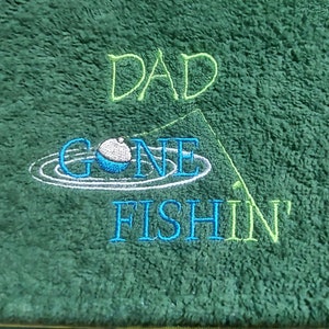 May include: A dark green textured towel featuring embroidered text that reads DAD GONE FISHIN'. The design includes a blue and white fishing bobber floating on water ripples and a thin green line representing a fishing rod and string.