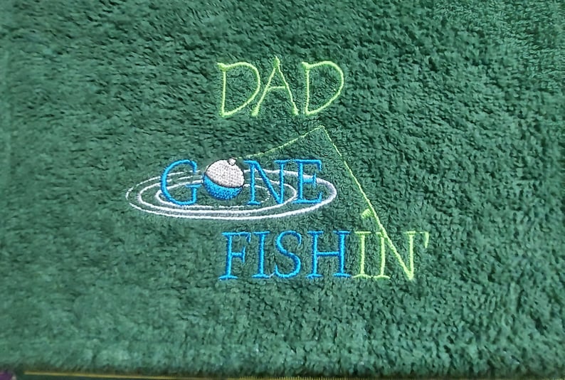 May include: A dark green textured towel featuring embroidered text that reads DAD GONE FISHIN'. The design includes a blue and white fishing bobber floating on water ripples and a thin green line representing a fishing rod and string.