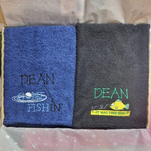 May include: Two folded rectangular towels. The blue towel features black embroidered text reading DEAN GONE FISHIN' with a fishing bobber graphic. The black towel features green text reading DEAN and IT WAS THIS BIG with a yellow fish graphic.