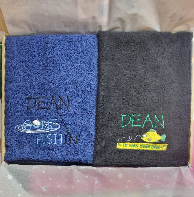 May include: Two folded rectangular towels. The blue towel features black embroidered text reading DEAN GONE FISHIN' with a fishing bobber graphic. The black towel features green text reading DEAN and IT WAS THIS BIG with a yellow fish graphic.