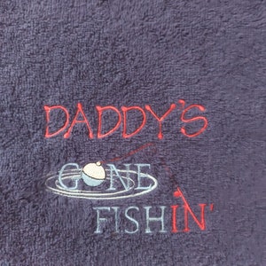 May include: A dark purple terry cloth towel featuring embroidered text that reads DADDY'S GONE FISHIN' in red and light blue thread. The design includes a stitched graphic of a fishing bobber floating on water ripples with a thin red fishing line.