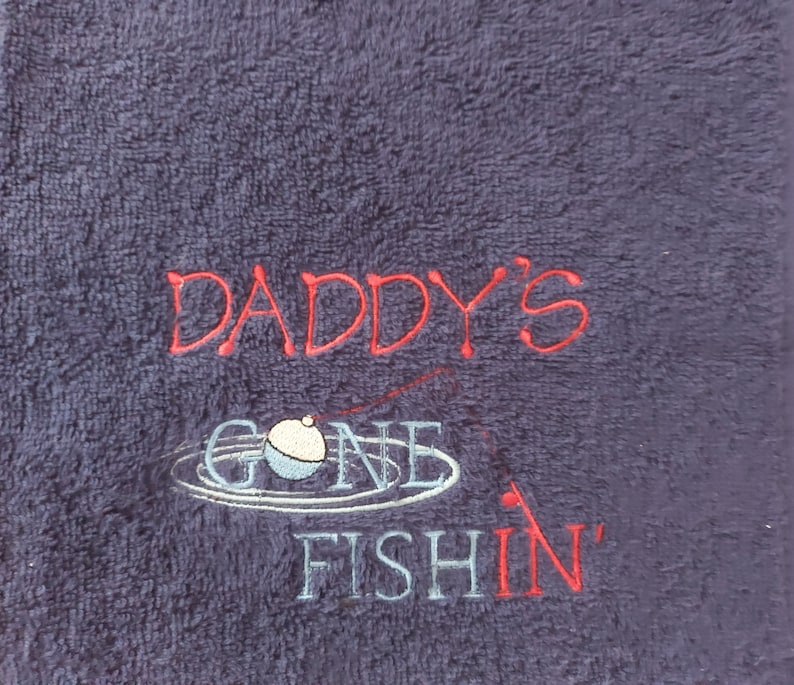May include: A dark purple terry cloth towel featuring embroidered text that reads DADDY'S GONE FISHIN' in red and light blue thread. The design includes a stitched graphic of a fishing bobber floating on water ripples with a thin red fishing line.