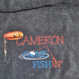 May include: A dark gray towel featuring embroidered red text that reads CAMERON and blue text that reads GONE FISHIN' with a fishing rod and bobber graphic. A metal fishing lure with an orange oval tag labeled CAMERON is attached to the towel.