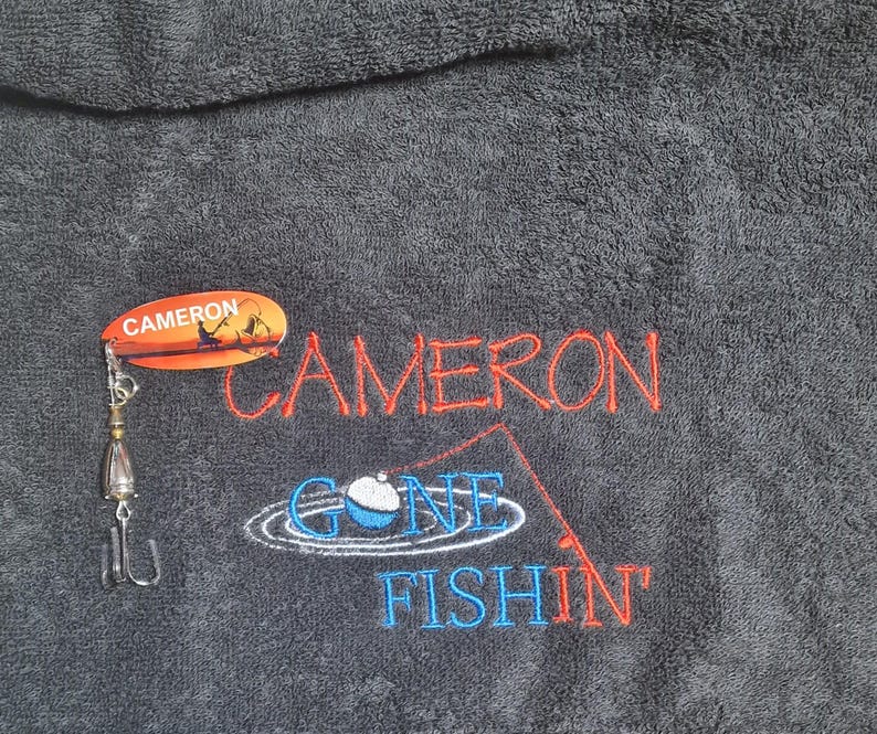 May include: A dark gray towel featuring embroidered red text that reads CAMERON and blue text that reads GONE FISHIN' with a fishing rod and bobber graphic. A metal fishing lure with an orange oval tag labeled CAMERON is attached to the towel.