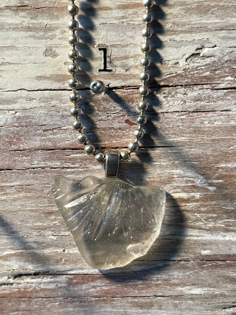 May include: A silver chain necklace with a clear glass pendant. The pendant is shaped like a seashell and has a textured surface.