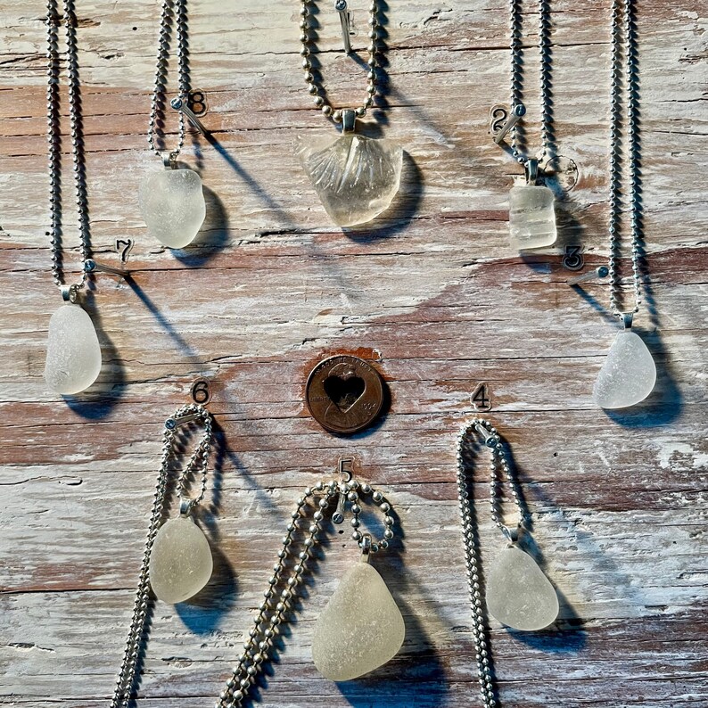 May include: Ten silver necklaces with various pendants, including sea glass and a shell, displayed on a white wooden background.