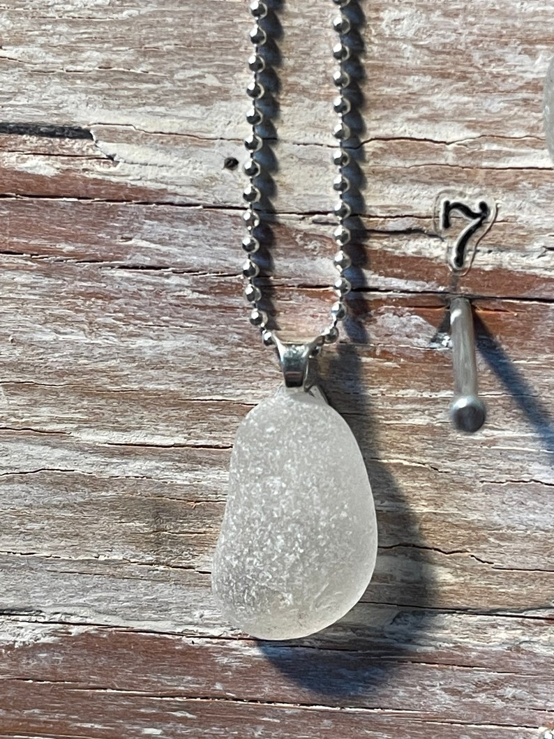 May include: A white sea glass pendant on a silver chain necklace. The pendant is a smooth, oval shape with a frosted finish.