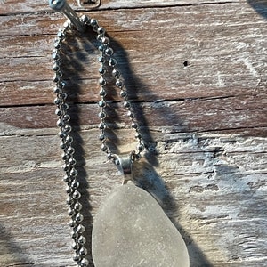 May include: A silver chain necklace with a white sea glass pendant. The necklace is hanging on a nail with the number "4" written on it.