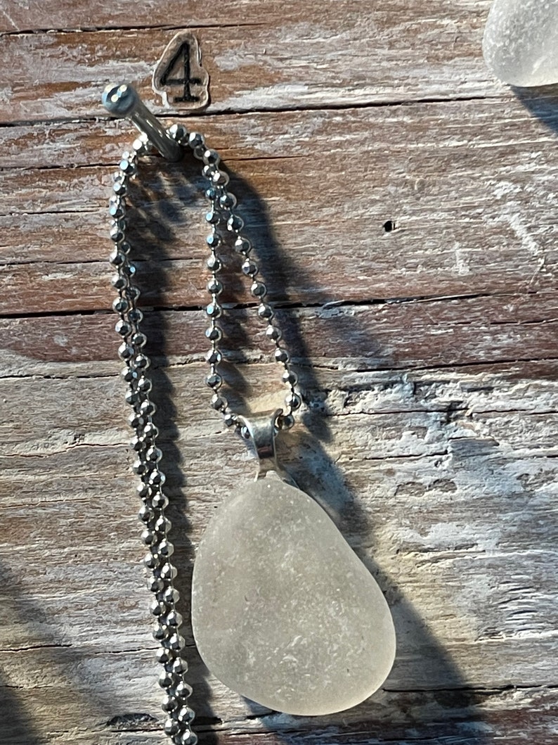 May include: A silver chain necklace with a white sea glass pendant. The necklace is hanging on a nail with the number "4" written on it.
