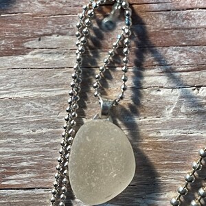 May include: A white sea glass pendant on a silver chain. The pendant is oval shaped and has a smooth, frosted surface.