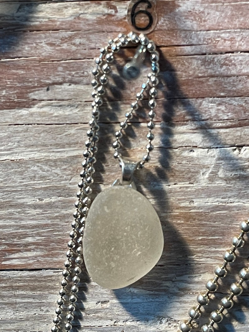 May include: A white sea glass pendant on a silver chain. The pendant is oval shaped and has a smooth, frosted surface.