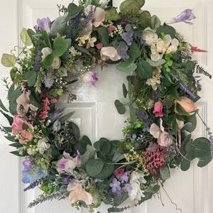 May include: A floral wreath with a variety of flowers and greenery. The wreath features pink, purple, white, and green elements. The wreath is circular and appears to be handmade, suitable for door or wall decor.