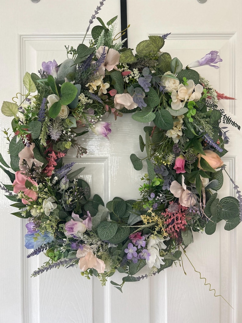 May include: A floral wreath with a variety of flowers and greenery. The wreath features pink, purple, white, and green elements. The wreath is circular and appears to be handmade, suitable for door or wall decor.