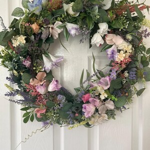 May include: A vibrant floral wreath featuring a variety of colorful flowers and greenery. The wreath includes white, pink, purple, and blue flowers, with green leaves. The wreath is a circular shape and is likely used for home decor.