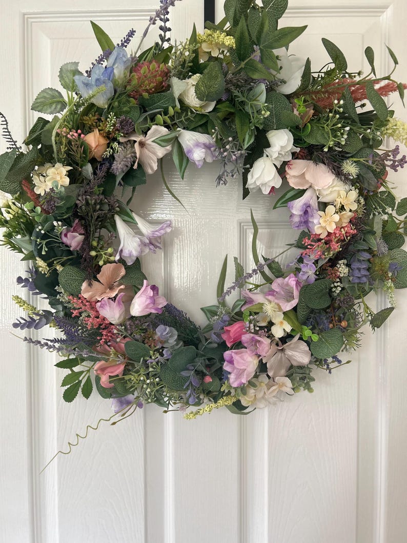 May include: A vibrant floral wreath featuring a variety of colorful flowers and greenery. The wreath includes white, pink, purple, and blue flowers, with green leaves. The wreath is a circular shape and is likely used for home decor.