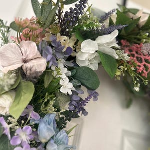 May include: A close-up of a floral wreath featuring a variety of artificial flowers in shades of pink, purple, white, and blue. Green leaves and sprigs of lavender are interspersed throughout the arrangement. The wreath is likely for home decor.