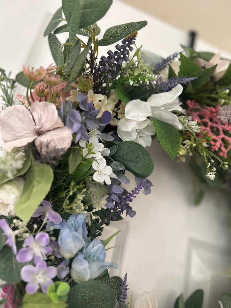 May include: A close-up of a floral wreath featuring a variety of artificial flowers in shades of pink, purple, white, and blue. Green leaves and sprigs of lavender are interspersed throughout the arrangement. The wreath is likely for home decor.