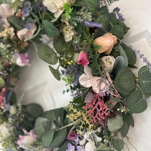 May include: A vibrant floral wreath featuring a variety of artificial flowers and greenery. The wreath includes pink, peach, white, and purple flowers, with green eucalyptus leaves. The wreath is a decorative item for doors or walls.