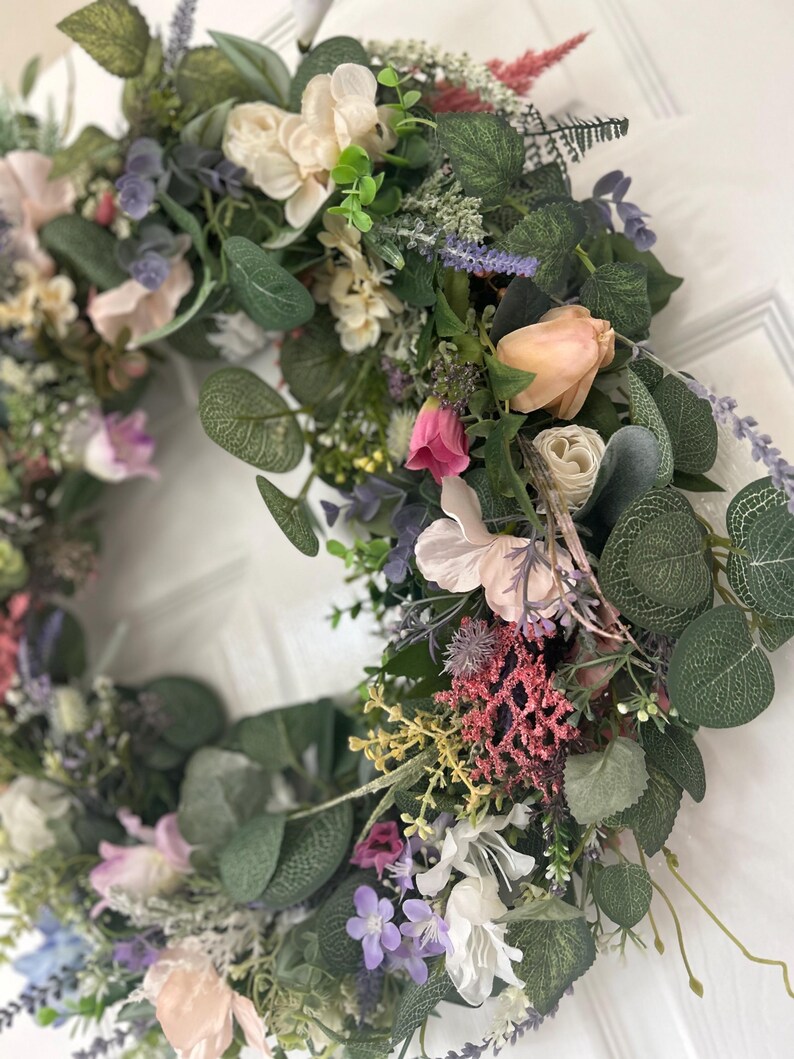 May include: A vibrant floral wreath featuring a variety of artificial flowers and greenery. The wreath includes pink, peach, white, and purple flowers, with green eucalyptus leaves. The wreath is a decorative item for doors or walls.