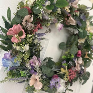 May include: A floral wreath with a variety of colorful flowers and green leaves. The wreath features pink, purple, white, and blue flowers, with green foliage. The wreath is likely used for home decor, such as a door decoration.