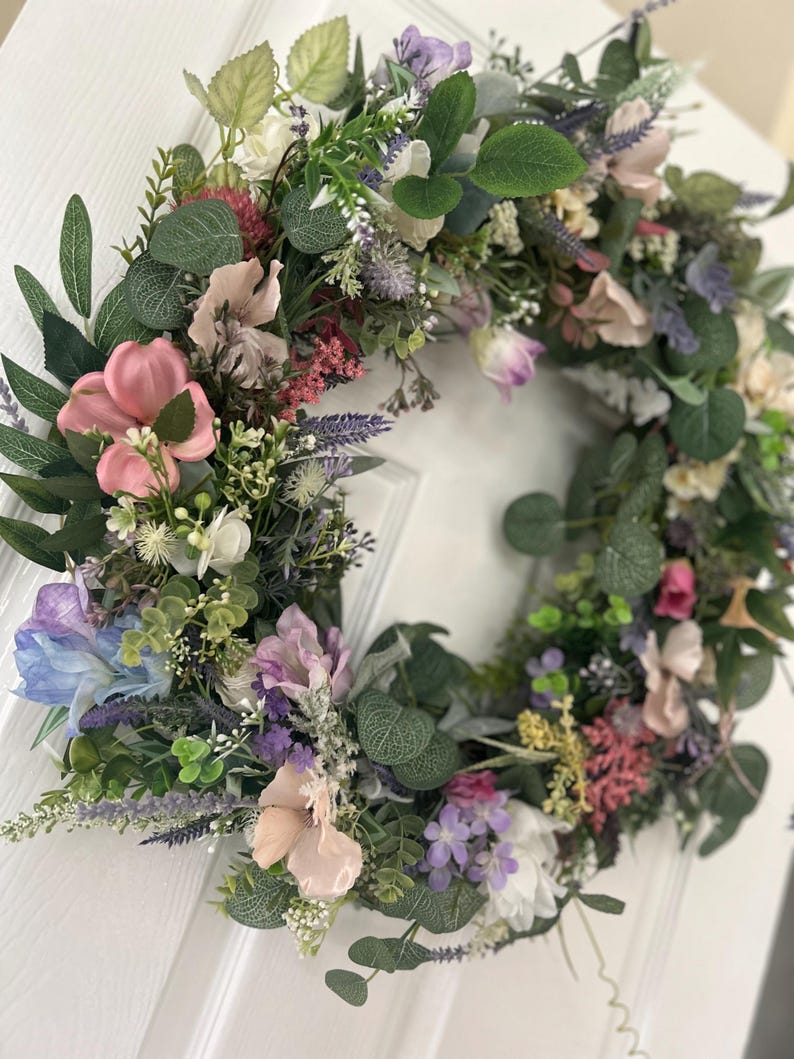 May include: A floral wreath with a variety of colorful flowers and green leaves. The wreath features pink, purple, white, and blue flowers, with green foliage. The wreath is likely used for home decor, such as a door decoration.
