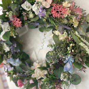May include: A vibrant floral wreath featuring a variety of colorful flowers and greenery. The wreath includes pink, white, purple, and blue flowers, with green leaves and stems. The wreath is likely used for home decor.