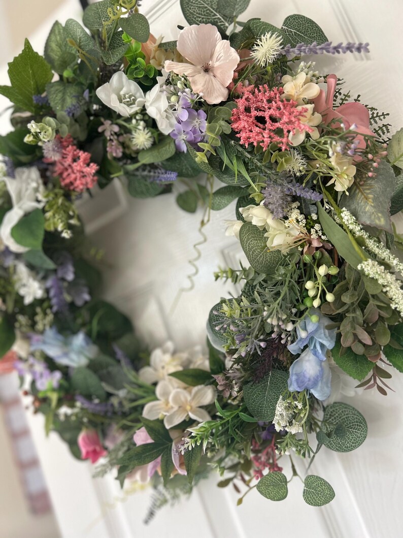 May include: A vibrant floral wreath featuring a variety of colorful flowers and greenery. The wreath includes pink, white, purple, and blue flowers, with green leaves and stems. The wreath is likely used for home decor.