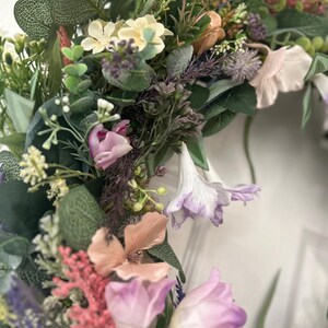 May include: A close-up of a floral wreath featuring a variety of flowers in shades of pink, purple, peach, and white, with green foliage. The wreath is densely packed with different types of flowers and greenery, creating a full and textured appearance.