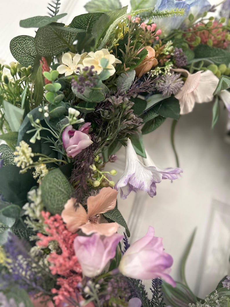 May include: A close-up of a floral wreath featuring a variety of flowers in shades of pink, purple, peach, and white, with green foliage. The wreath is densely packed with different types of flowers and greenery, creating a full and textured appearance.