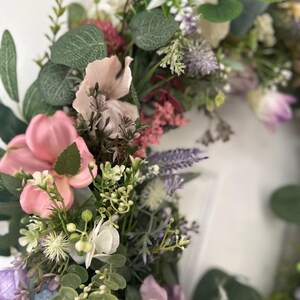 May include: A close-up of a floral wreath featuring a variety of artificial flowers in shades of pink, purple, white, and green. The wreath includes large pink blossoms, lavender sprigs, and green foliage, creating a vibrant and textured display.