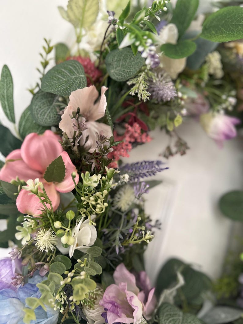 May include: A close-up of a floral wreath featuring a variety of artificial flowers in shades of pink, purple, white, and green. The wreath includes large pink blossoms, lavender sprigs, and green foliage, creating a vibrant and textured display.