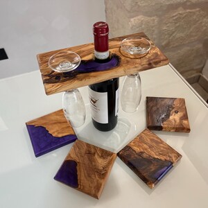 Può includere: Un porta bottiglie in legno con due porta bicchieri e una bottiglia di vino. Il porta bottiglie è in legno d'ulivo e ha un'intarsio in resina epossidica viola. Ci sono quattro sottobicchieri con lo stesso design.