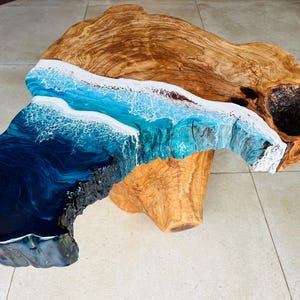 May include: A unique, handcrafted coffee table. The table features a natural wood top with a striking epoxy resin design. The resin mimics ocean waves in shades of blue and white, creating a coastal aesthetic. The table's dimensions are approximately 48 inches long, 30 inches wide, and 18 inches high.